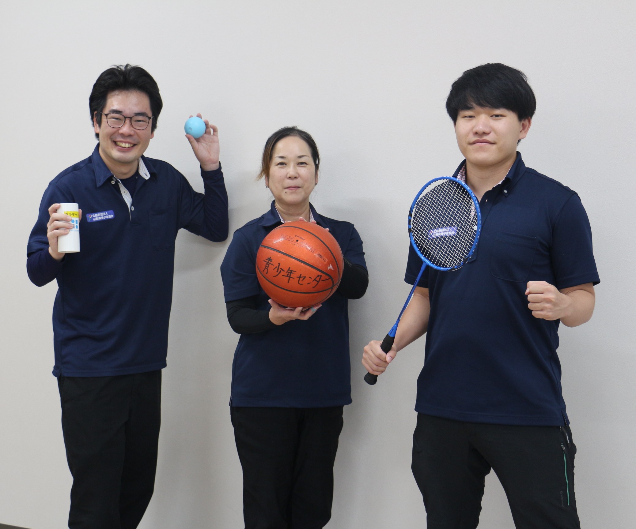 体験活動で使用するスポーツ用品を持っているスタッフ、左からジャグリング道具(筒けん)を持っている男性スタッフ、バスケットボールを持っている女性スタッフ、バトミントンラケットを持っている男性スタッフ